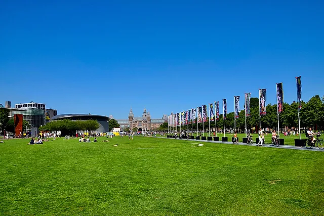 The wide lawn of Museumplein under a blue sky, with a row of flags and the Rijksmuseum and Van Gogh Museum on the horizon